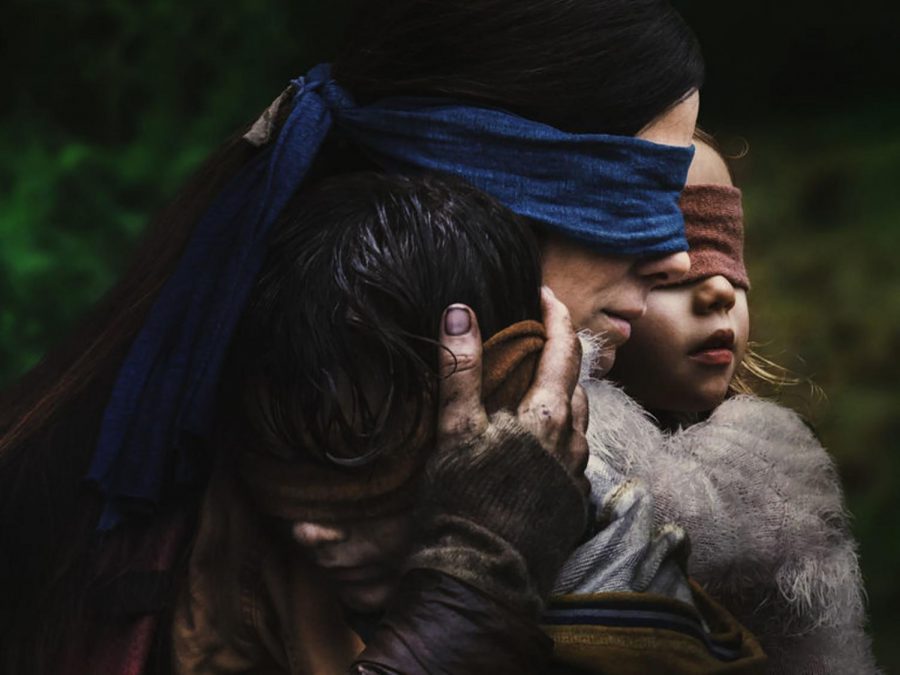 Malorie Hayes, played by Sandra Bullock, holds two children in her arms while wearing a blindfold. Bird Box is set in an apocalyptic world where a monster drives all who see it to kill themselves.