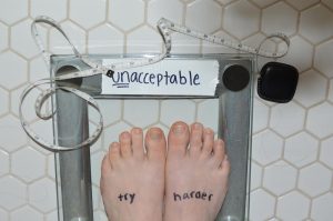 In this photo staged by junior Emily Eshel, a girl stands on a scale that reads unacceptable where the weight is supposed to be located. Eshel created this image for an English project exploring beauty ideals. Sophomore Leah Abazari said that the pressure to be a certain weight and a certain body type can be overwhelming.