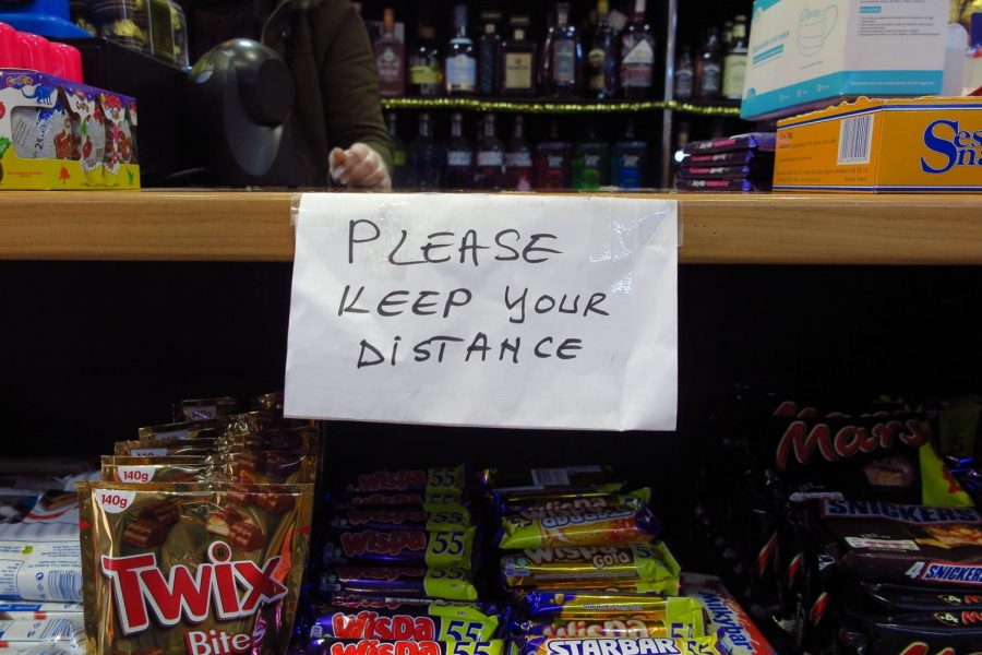 A liquor store in Sheffield, UK posts a sign reminding customers to social distance during the COVID-19 pandemic. Although businesses that provide essential services like food or healthcare are permitted to stay open, millions of people, including members of the Archer community, face financial uncertainty and job insecurity. 