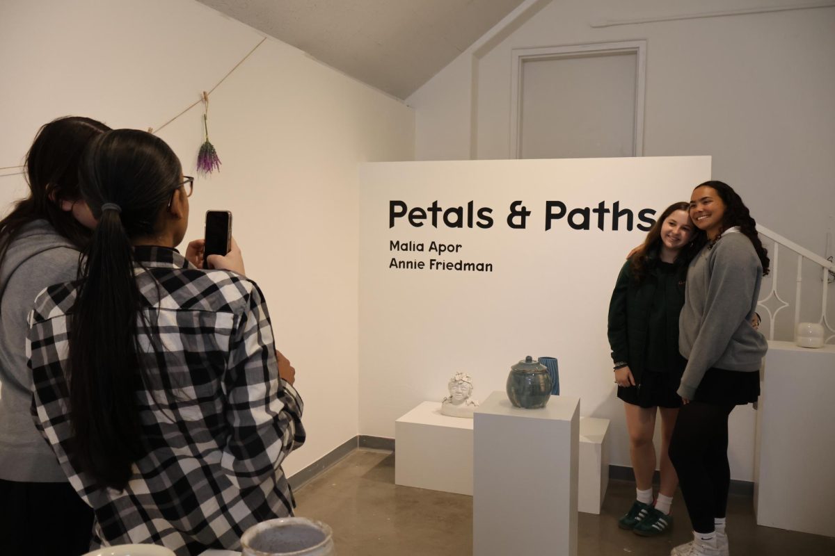 Annie Friedman (’24) and Malia Apor (’24) pose for a photo in front of the title wall for their Petals and Paths senior art show. Friedman and Apor are Advanced Study Ceramics students and chose to display pottery with themes of nature and family Wednesday, March 6. “I’m drawn to using one glaze called overglaze to make it more cohesive,” Friedman said. “I know Malia uses something called underglazing sometimes, and it’s cool that there’s multiple different ways.” 