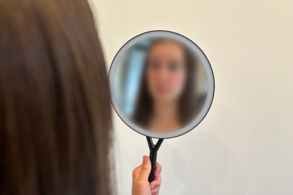 I hold a handheld mirror, looking at my blurred reflection. Society has conditioned women to feel unsatisfied with their appearance. Photo illustration by Phoebe Measer.