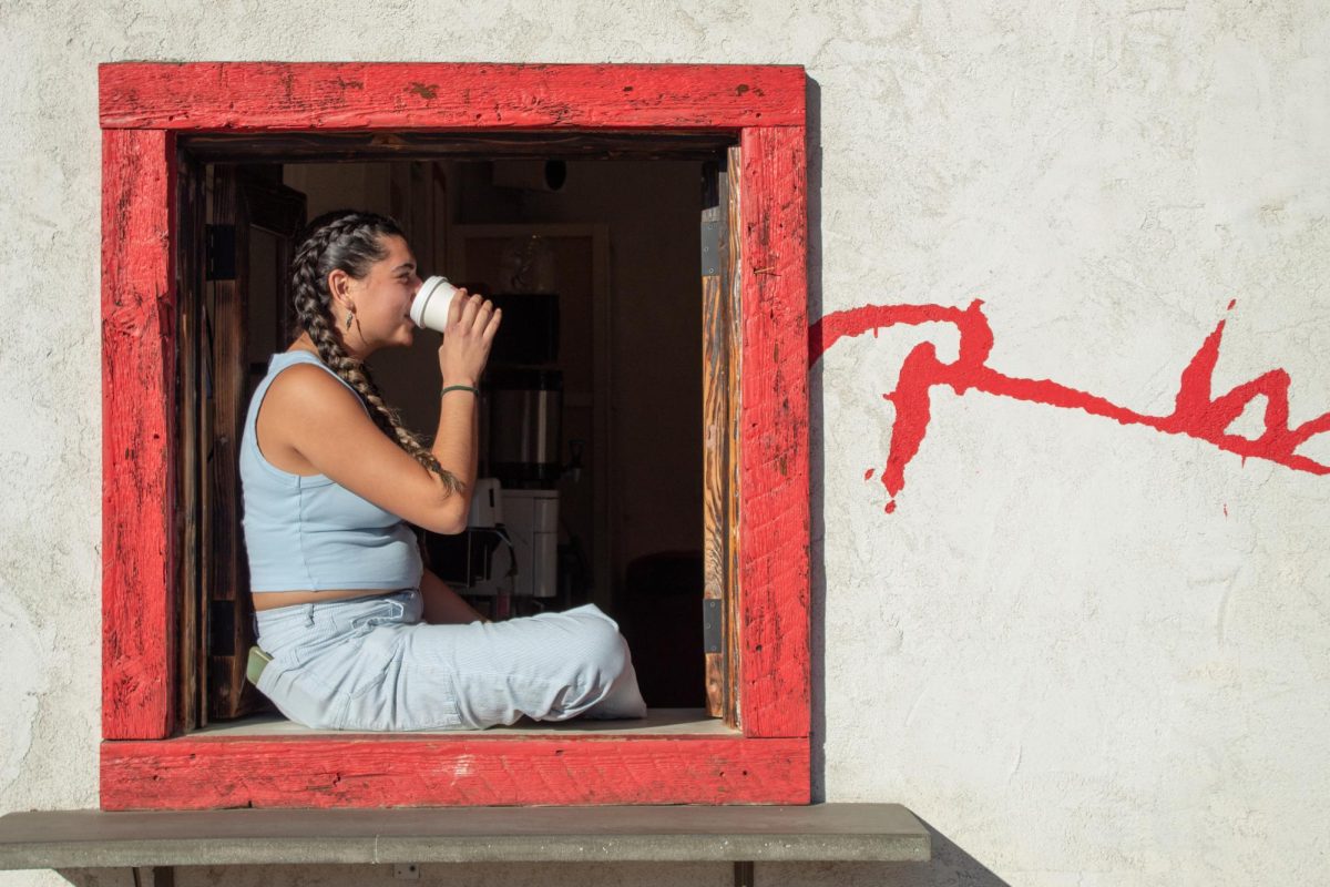 A Red Window barista sits in the frame of their iconic red window, drinking a warm drink. From handing drinks through the window, sitting on the sill or even peeking inside to see baristas hard at work, the red window gives Red Window a distinct feel.