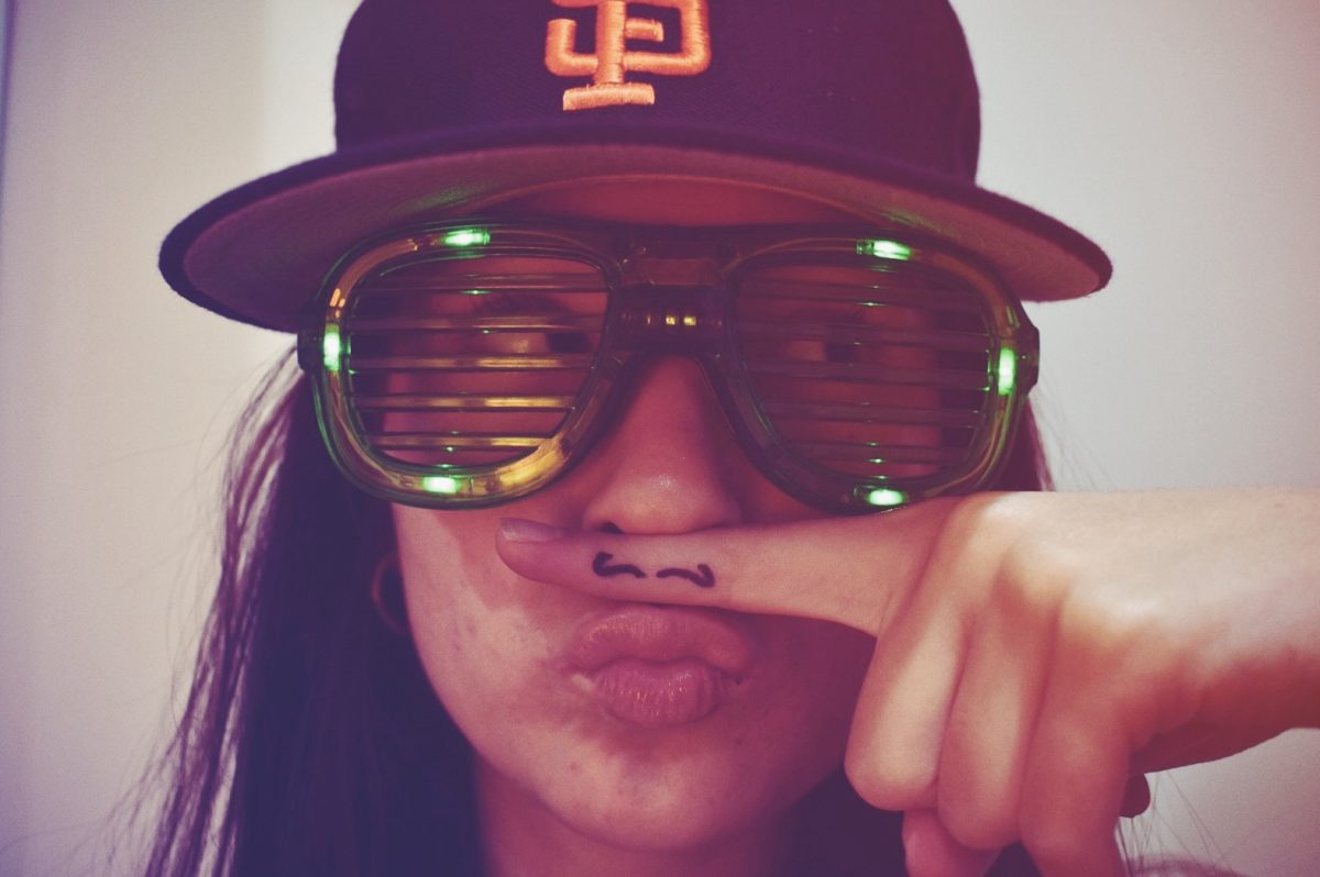 A person poses in shutter shades and a snapback, holding a finger with a drawn on mustache up to their face. 2016 internet culture often included these fashion pieces in social media posts. The finger mustache was popular on Tumblr, Vine and early Instagram. 